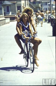 two women riding bikes down the street on a sunny day with people in the background