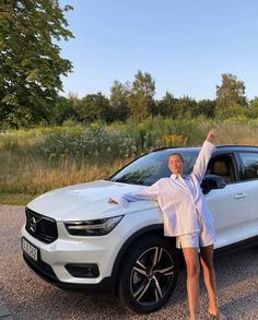 a woman standing in front of a white car with her arms up and hands raised