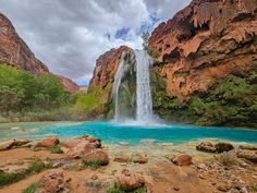 the waterfall is surrounded by large rocks and blue water in front of red cliffs with green trees on either side