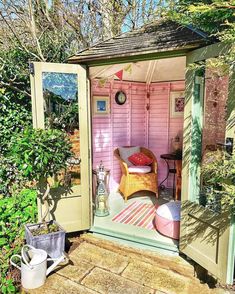 a small pink shed with a chair and table in the doorway, surrounded by greenery