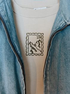 a blue jean jacket with a mountain stamp on the front and back pocket, hanging from a white t - shirt