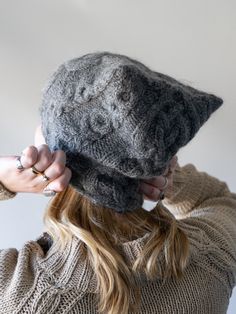 a woman wearing a gray knitted hat while holding her hair in one hand and looking at the camera