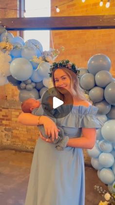 a woman in a blue dress holding a baby while standing next to balloons and flowers