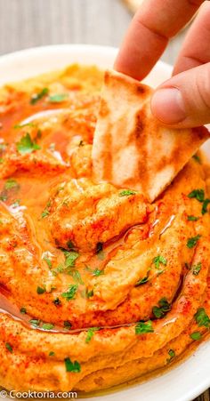 a hand dipping a tortilla chip into a bowl of hummus and sauce