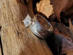 two wedding rings sitting on top of a piece of wood with leaves around them and one ring has a princess cut diamond in the middle