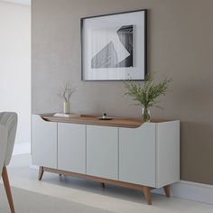 a white sideboard with two plants on it in front of a chair and framed photograph