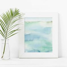 a watercolor painting in a white frame next to a vase with a green plant