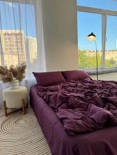 an unmade bed with purple sheets and pillows in front of a large window overlooking the city