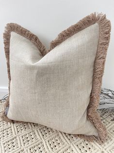 a white pillow with brown fringe on top of a woven table cloth area next to a wall