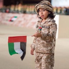 a little boy in uniform holding a flag and pointing to it with one hand while standing on the ground