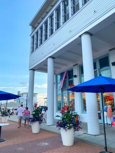 people walking on the sidewalk in front of a building with blue umbrellas and potted plants