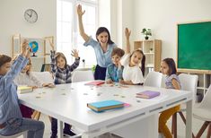 a group of children sitting at a table with their arms in the air and two adults standing behind them