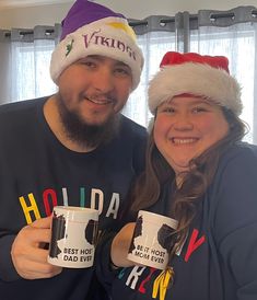 a man and woman holding coffee mugs in front of a christmas hat on their heads