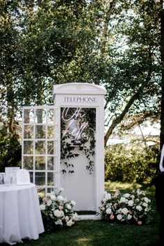 a white phone booth with flowers and greenery on the grass next to a table