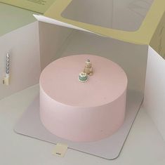 a pink cake in a box with a small figurine sitting on top of it