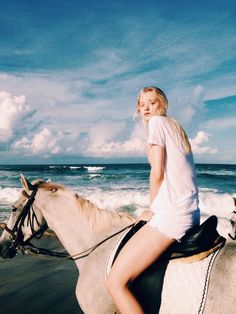 a woman riding on the back of a white horse down a beach next to the ocean