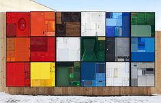 a wall made up of many different colored doors in snow covered ground next to building