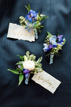 three boutonnieres with blue flowers and greenery are on a black cloth