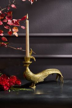 a candle holder with a gold rooster on it next to red flowers and a branch