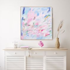 an abstract painting hangs on the wall above a white dresser with drawers and vases