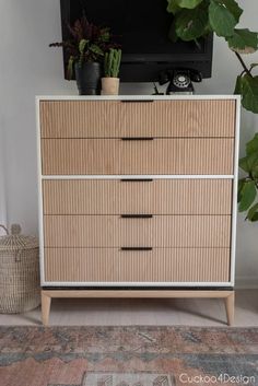 a white dresser with brown drawers and plants on the top, in front of a flat screen tv