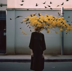 a woman standing on the street with birds flying over her head and yellow flowers in front of her