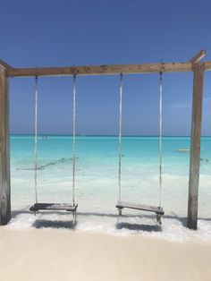 two wooden swings sitting on top of a sandy beach next to the ocean with clear blue water