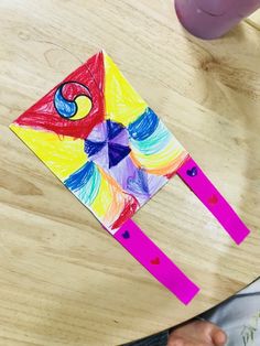 a child's hand holding a colorful kite on top of a wooden table next to a purple cup