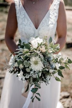 a woman in a white wedding dress holding a bouquet with greenery and flowers on it