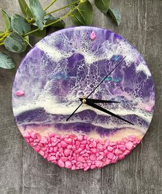 a clock with pink and purple pebbles on it sitting next to a plant in front of a wooden surface