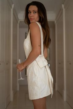 a woman in a white dress holding a wine glass and looking at the camera while standing in a hallway