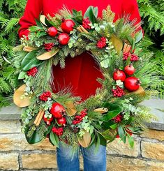 a person standing in front of a brick wall holding a wreath with red berries and greenery