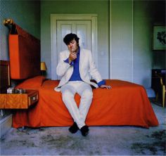 a man sitting on top of a bed next to an orange suitcase in a room