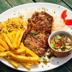 a white plate topped with steak and french fries next to a bowl of sauce on top of rice