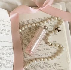 an open book with pearls and a bottle on it next to a pink ribbon tied around the book