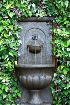 a water fountain surrounded by greenery in front of a wall with flowers and leaves