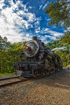 an old fashioned steam engine train on the tracks near some trees and people walking around