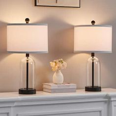 two lamps sitting on top of a white mantle next to a book and vase with flowers