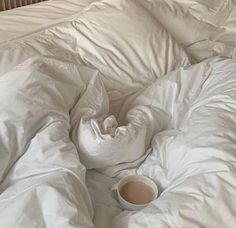 an unmade bed with white sheets and a cup of coffee on top of it