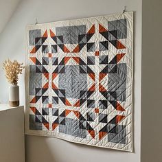 a quilt hanging on the wall next to a potted plant and vase with dried flowers