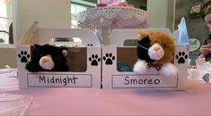 two stuffed animals are sitting in small boxes on a pink table cloth covered tablecloth