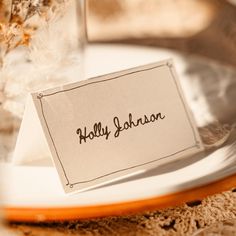 a place card sitting on top of a white plate next to a glass filled with flowers