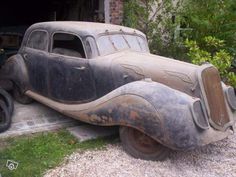 an old, rusty car is parked in front of a building with trees and bushes