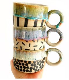 a hand holding a stack of coffee mugs in different colors and designs on them