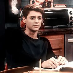 a young man sitting at a table with a book and pen in his hand while looking at the camera