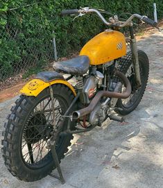 an old yellow motorcycle parked on the sidewalk next to a chain link fence and shrubbery