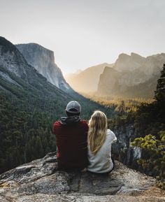 two people sitting on top of a cliff looking at the mountains and trees in the distance