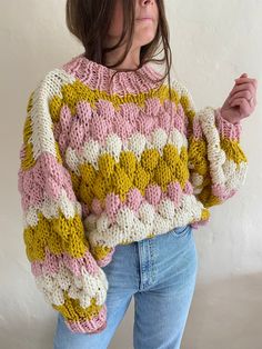 a woman wearing a pink, yellow and white knitted sweater standing in front of a wall