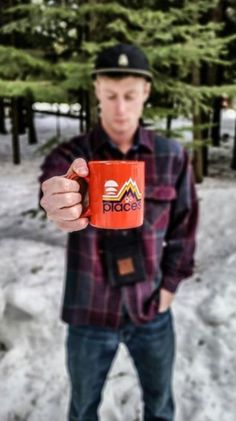 a man holding up a red coffee mug in front of snow covered ground and trees