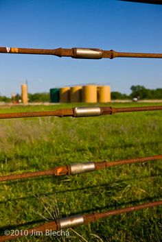 an old rusty fence in the middle of a grassy field with grain silos in the background